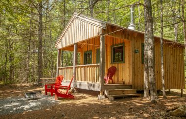 A wooden Kejimkujik camping lodge in the woods with red chairs on the deck