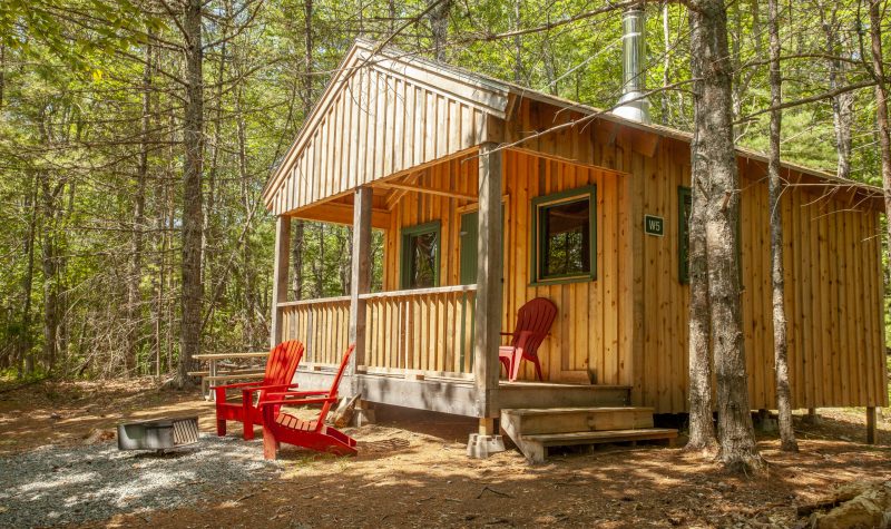 A wooden Kejimkujik camping lodge in the woods with red chairs on the deck