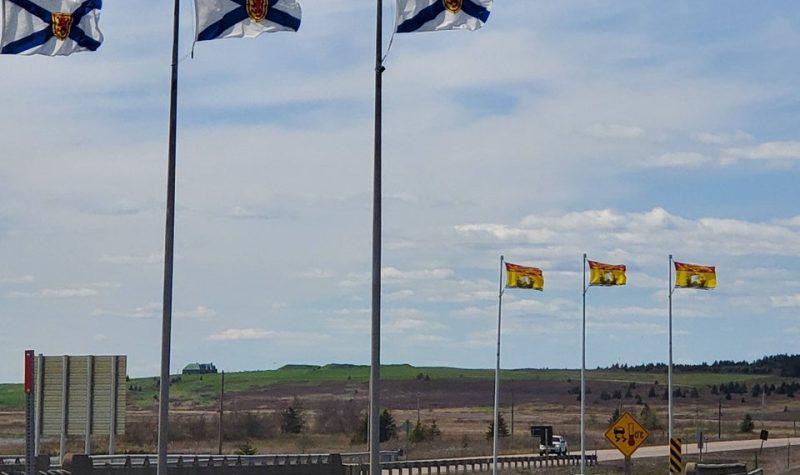 Three Nova Scotia flags on poles in foreground, and three New Brunswick flags on poles in the background, along a highway.