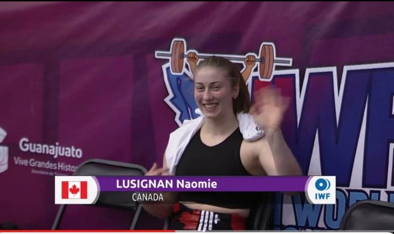 Naomie Lusignan waves at the camera wearing a weightlifting singlet in front of a purple background.