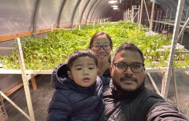 A man, woman and young child pose in a greenhouse with young green plants growing behind them.