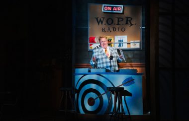 A man in a bright jacket in a spotlight speaks under an on-air setup in a play.