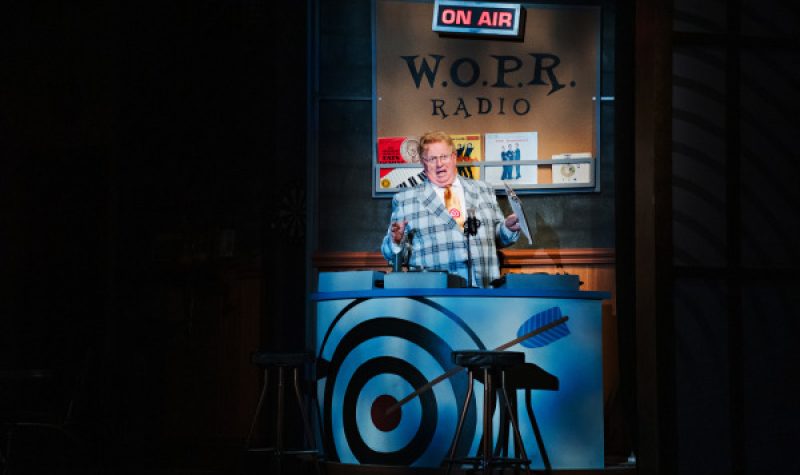 A man in a bright jacket in a spotlight speaks under an on-air setup in a play.