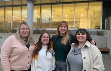 Four women, stand smiling in front of a building