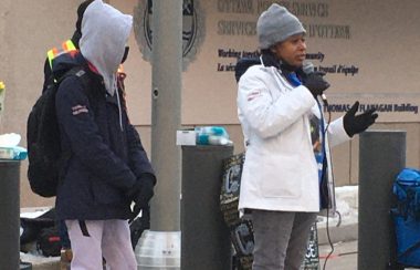 A woman wearing a white winter coat, grey toque, and black boots speaks to an outdoor crowd through a microphone.
