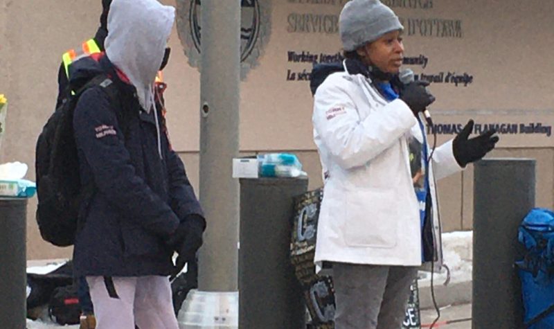 A woman wearing a white winter coat, grey toque, and black boots speaks to an outdoor crowd through a microphone.