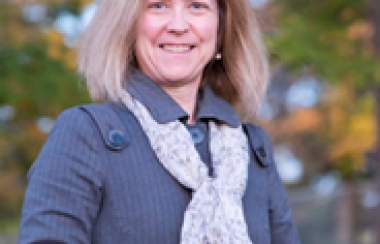 A shot of Nicole Thompson standing in front of trees in autumn wearing a grey coat and a white scarf.