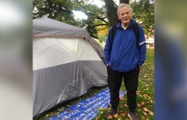 Phil, an encampment resident at Allan Gardens Park, stands next to a sign behind his tent which reads “It is not over, it’s only just beginning. It’s not about revolution, it’s about solutions and becoming one again. It’s a new beginning. Start your own community, build your own home.”