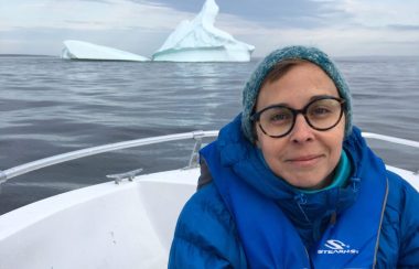 Une femme habillée chaudement pose devant un iceberge au loin. On ne voit qu'une partie du bateau dans lequel elle se trouve.