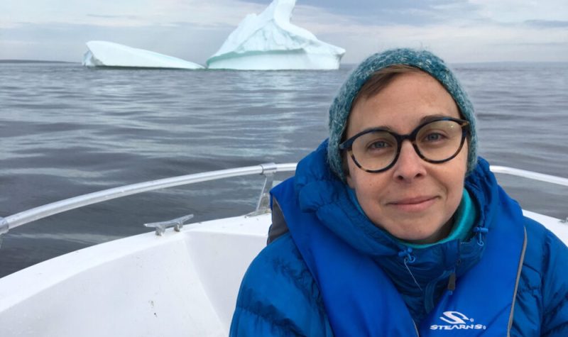 Une femme habillée chaudement pose devant un iceberge au loin. On ne voit qu'une partie du bateau dans lequel elle se trouve.