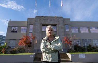 A woman wearing a light green jacket stands in front of Nanaimo City Hall with her arms crossed