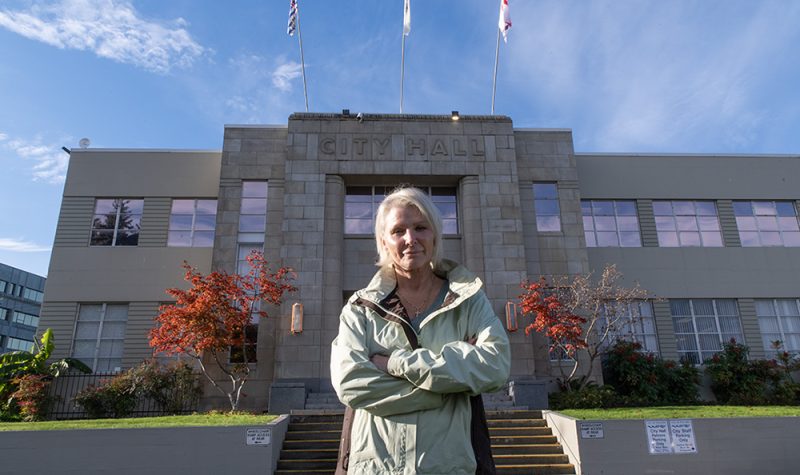 A woman wearing a light green jacket stands in front of Nanaimo City Hall with her arms crossed