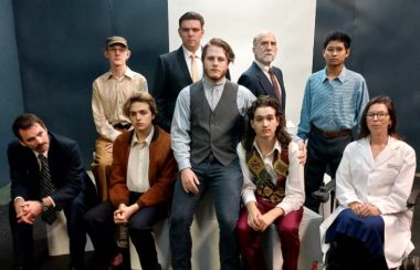 A group of actors are shown sitting and standing in two rows, wearing costume and apparently in character..