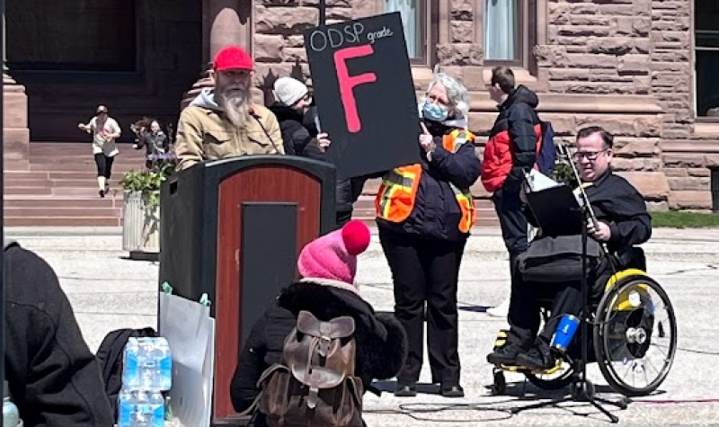 People at a podium speaking and holding up a sign with the letter F.