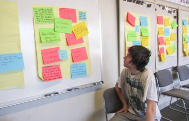 A man sits in front of a chalkboard looking at the papers being displayed on them. Different coloured pieces of papers are being displayed all on the walls of the classroom.