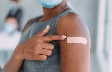 A person with dark skin, wearing a grey shirt and a white mask, points at a bandage on their arm, where we can assume they have just been vaccinated.