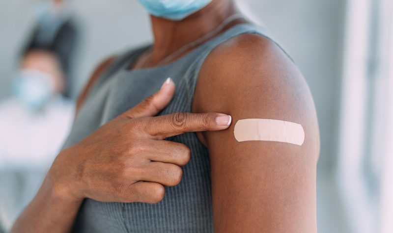 A person with dark skin, wearing a grey shirt and a white mask, points at a bandage on their arm, where we can assume they have just been vaccinated.