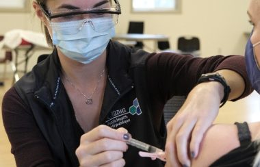 An Ottawa Public Health nurse wearing a medical mask, protective eye wear, and a black polo shirt administers a vaccine into the upper arm of a patient.