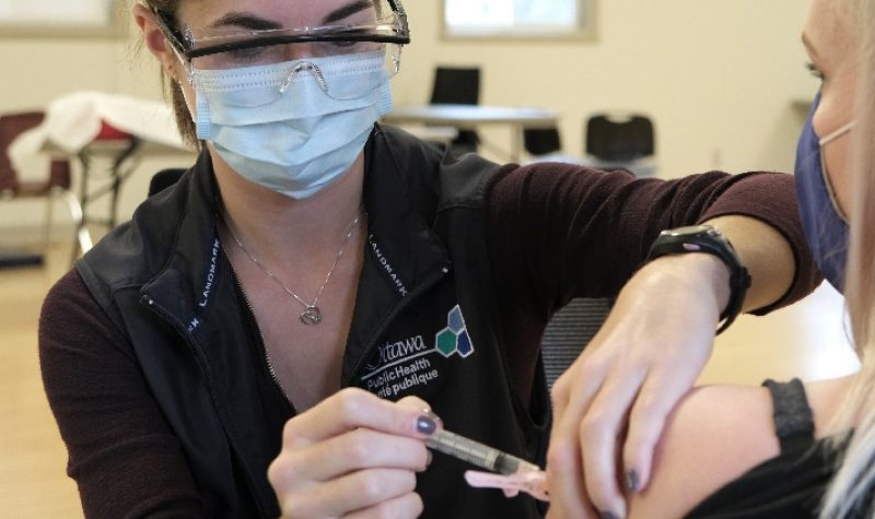 An Ottawa Public Health nurse wearing a medical mask, protective eye wear, and a black polo shirt administers a vaccine into the upper arm of a patient.