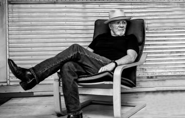 Une photo en noir et blanc, d’un homme assis sur une chaise posant une jambe sur l’autre et regardant à côté.