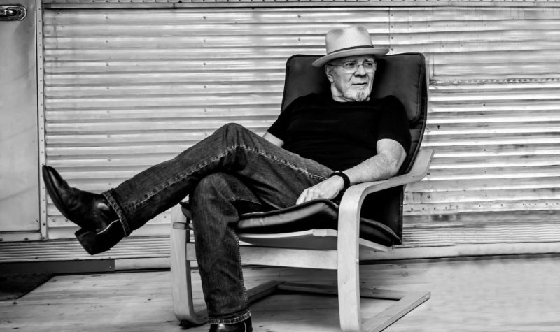 Une photo en noir et blanc, d’un homme assis sur une chaise posant une jambe sur l’autre et regardant à côté.