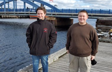 Olivia and Garfield Gallant-Zwicker stand side by side in front of a bridge over Mersey River on a sunny day