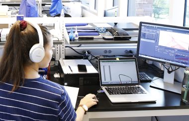 Student with headphones sits at computer screens and operates a mouse.