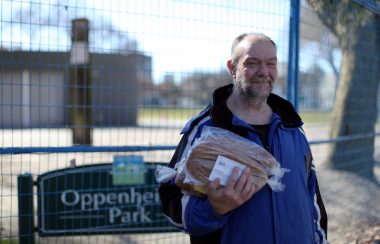 Downtown Eastside resident Paul Young holds loaves of bread outside with a green Oppenheimer Park sign behind him
