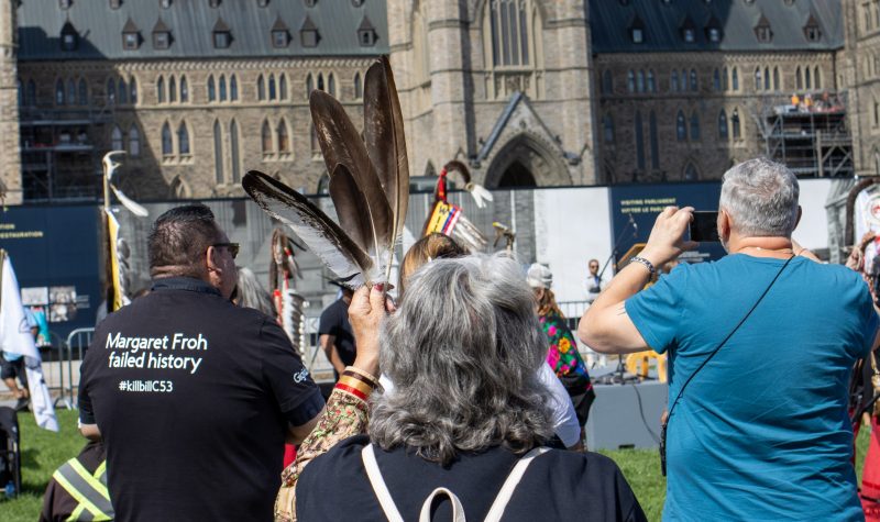 Three people face the House of Commons with their back to the camera. The woman in the centre is holding 3 feathers.