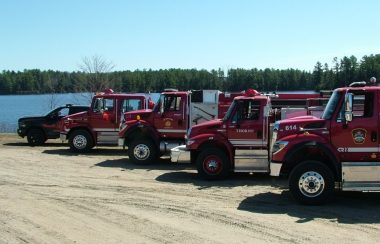 A photo of four red firetrucks and a black and red emergency vehicle lined up in a dirt parking lot next to a lake.