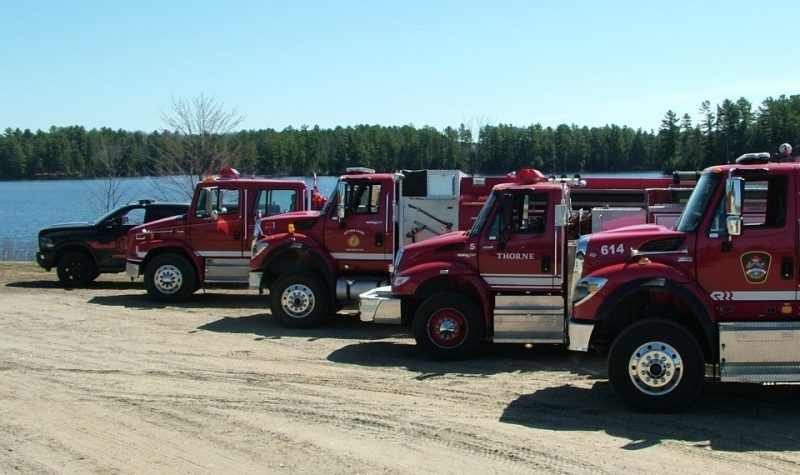 A photo of four red firetrucks and a black and red emergency vehicle lined up in a dirt parking lot next to a lake.