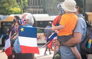 Un père porte ses enfants, ils tiennent un drapeau acadien, au fond il y a des personnes habillés aux couleurs de l'Abadie et un bus d'Halifax Transit