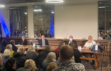 Four municipal political candidates sitting at a long table and facing a large group of seated people. Behind the candidates are mirrors reflecting the crowded room.