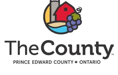 A logo featuring the words 'The County, Prince Edward County Ontario' underneath a graphic with a barn, grapes and water.