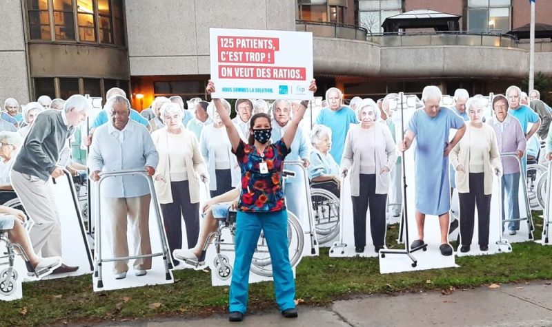 Une infirmière tenant une pancarte. Elle est debout devant plusieurs portraits de gens en carton grandeur nature.