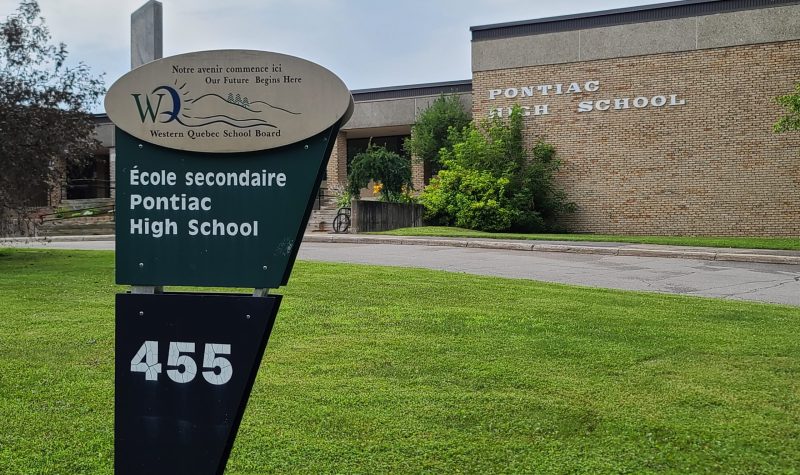 The exterior of a one story brown brick building with a sign reading Pontiac High School. There is a triangular sign on a green lawn in front of the building with the school's name in French and the number 455.