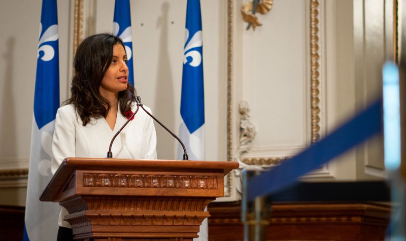 Dominique Anglade derrière un lutrin en bois, portant une blouse blanche et entouré de drapeaux du Québec