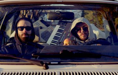 Two men wearing sunglasses sitting in a car with music instruments behind them, seen through the windshield.
