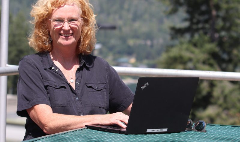 A woman sits in front of a computer outside. Trees in the background.