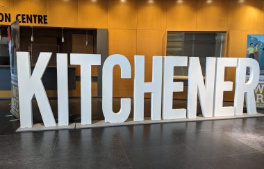 Large 3-feet tall letters spell out the word 'Kitchener' inside a building with a brown floor and orange coloured walls.