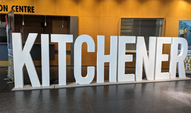 Large 3-feet tall letters spell out the word 'Kitchener' inside a building with a brown floor and orange coloured walls.