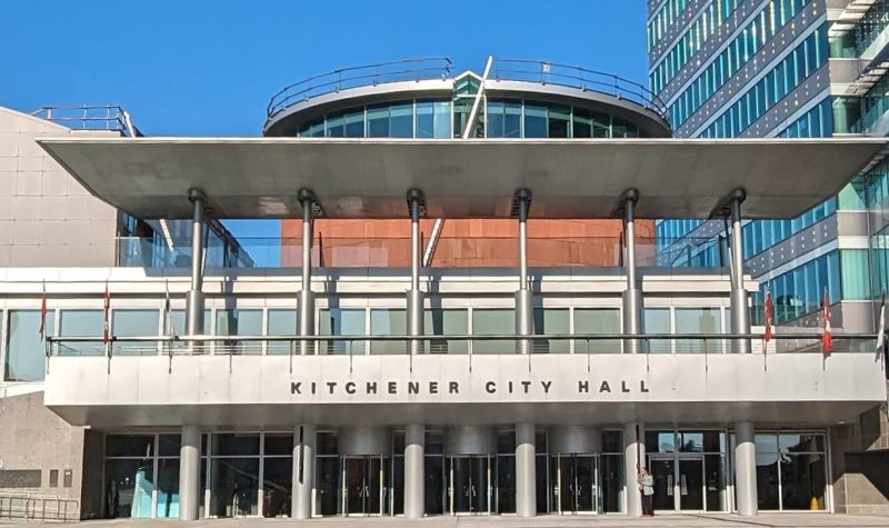 A picture of the front of a 2 story modern building with the words 'Kitchener City Hall' across the front of the first level.