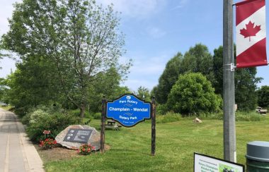 On peut voir le panneau bleu d'identification du parc Rotary Champlain-Wendat. Le gazon et les arbres du parc sont visibles en arrière-plan et une banderole aux couleurs du drapeau canadien est visible à proximité.