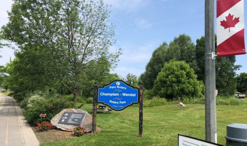 On peut voir le panneau bleu d'identification du parc Rotary Champlain-Wendat. Le gazon et les arbres du parc sont visibles en arrière-plan et une banderole aux couleurs du drapeau canadien est visible à proximité.