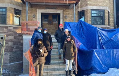 People in winter jackets standing on the steps of an apartment building outdoors with blue tarps on the right side.