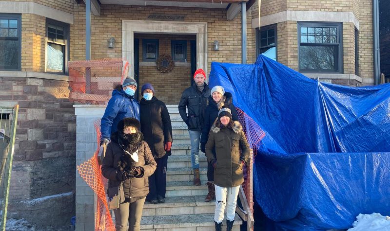 People in winter jackets standing on the steps of an apartment building outdoors with blue tarps on the right side.