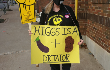 Woman holding two signs, a CUPE NB fist, and a homemade sign saying Higgs is a dictator.