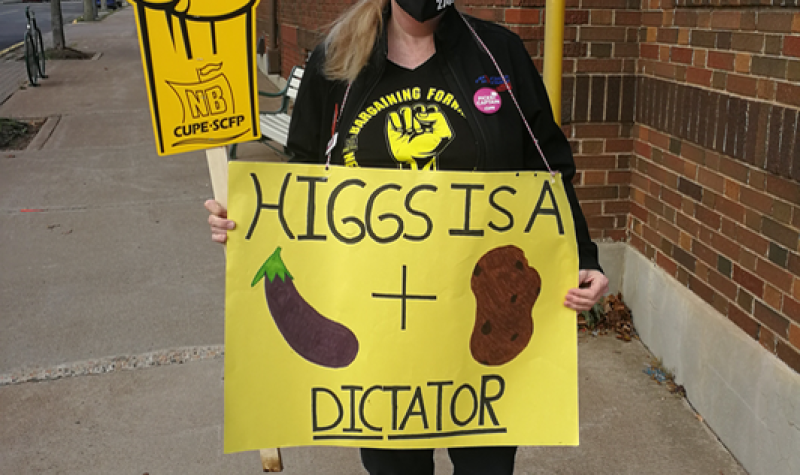 Woman holding two signs, a CUPE NB fist, and a homemade sign saying Higgs is a dictator.