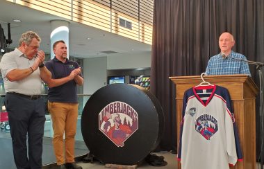 Two men applaud as another man speaks at a podium, unveiling new uniforms for the South Shore Junior B Lumberjacks hockey team