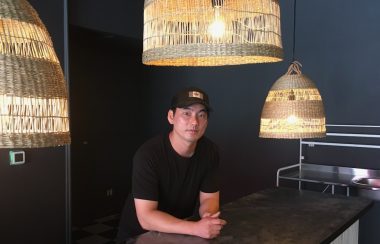 A young man in a ball cap leans on a black counter with rattan lighting hanging around him, and black walls behind him